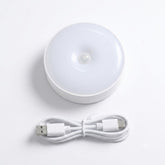 A white circular LED light with a built-in sensor, shown alongside a coiled white USB charging cable on a clean white background.