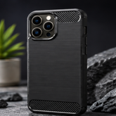 A black carbon-fiber-texture phone case with a raised camera bumper, shown standing upright on a dark surface with rocks and a small plant in the background.