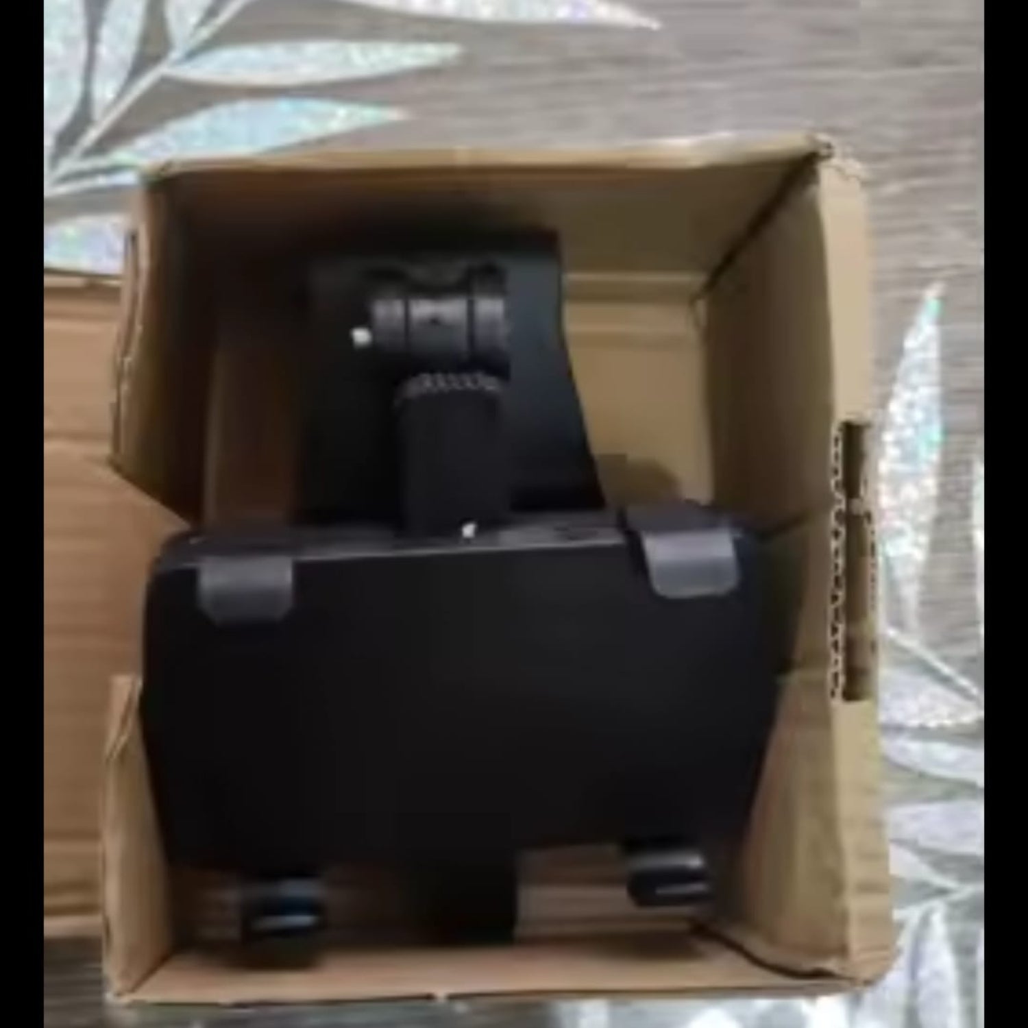 Black suitcase with wheels inside a cardboard box on a reflective surface.