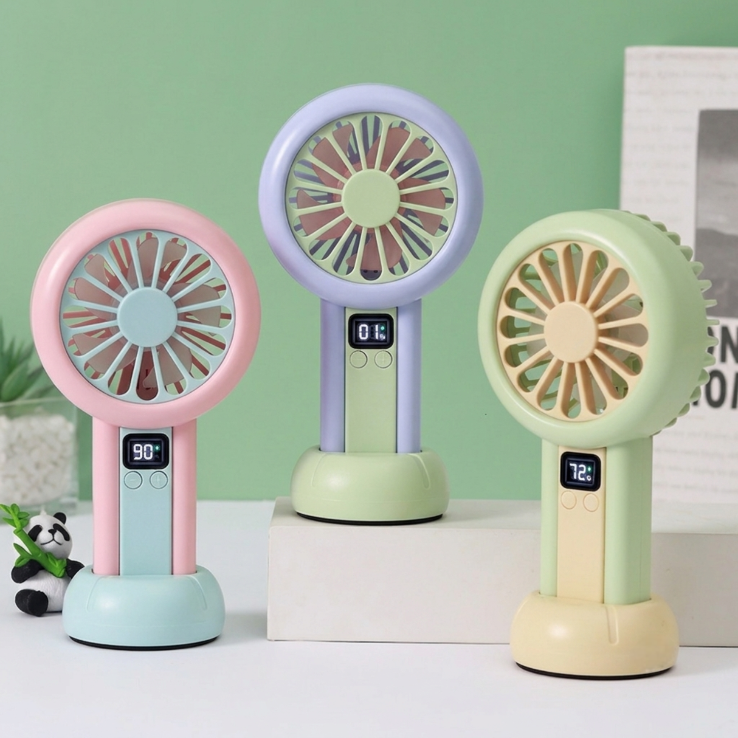 Three colorful handheld fans on a white surface with a green wall background.