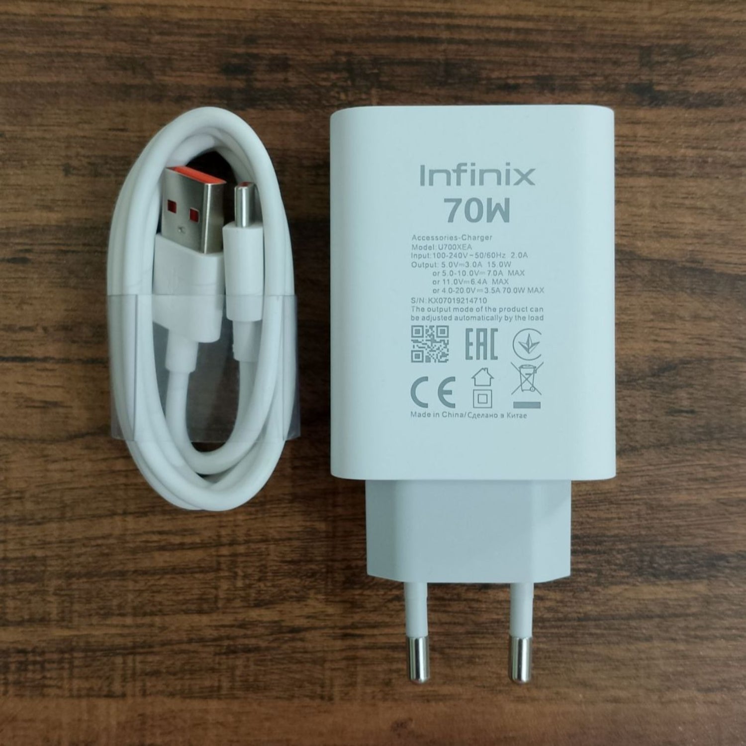 An Infinix 70W white wall charger adapter next to a white USB-C cable with a USB-A and USB-C connector, displayed on a dark wood background.