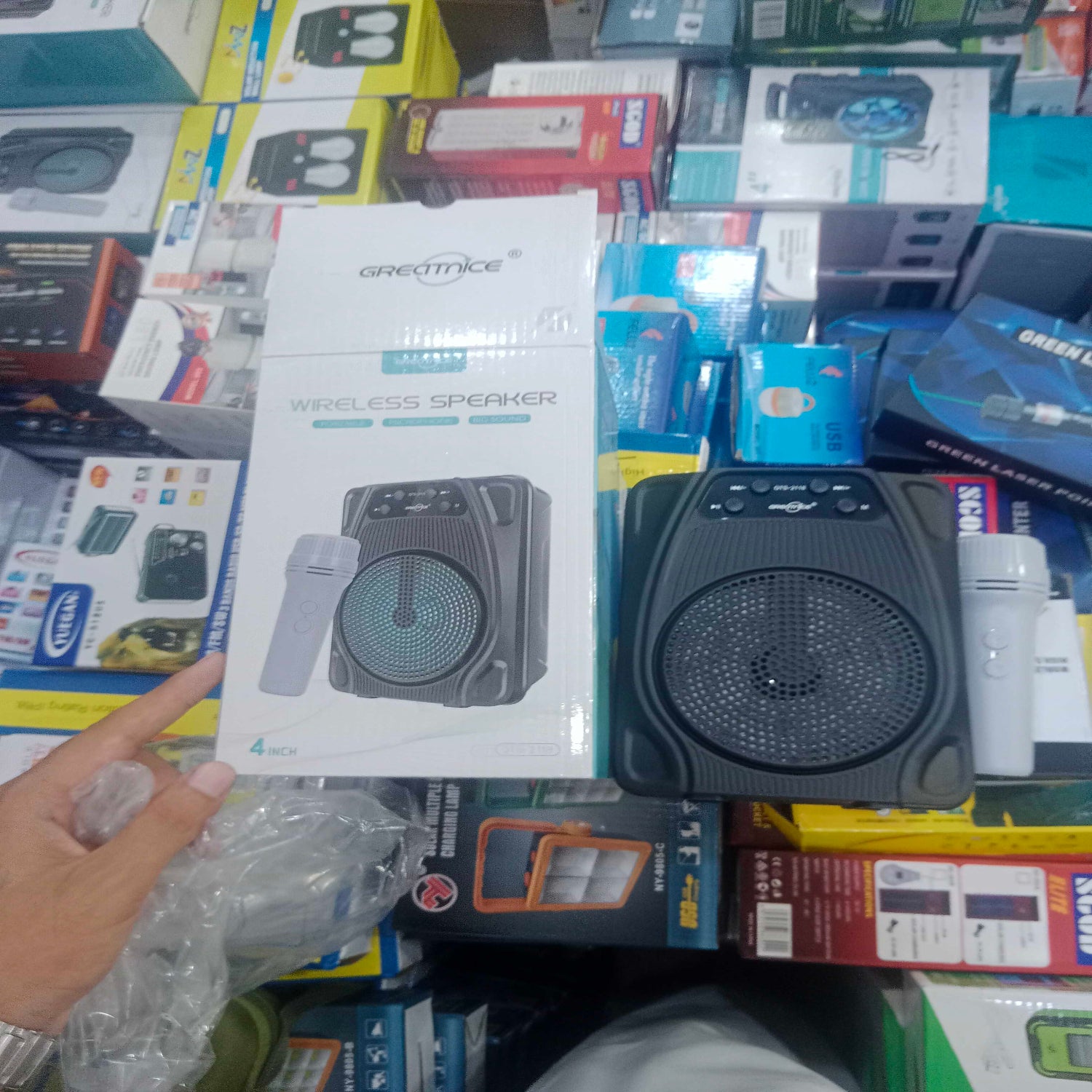 A retail store shelf display showing a Greatmice wireless speaker in its original white packaging box, alongside the black speaker itself, with a hand pointing at the box on the left.
