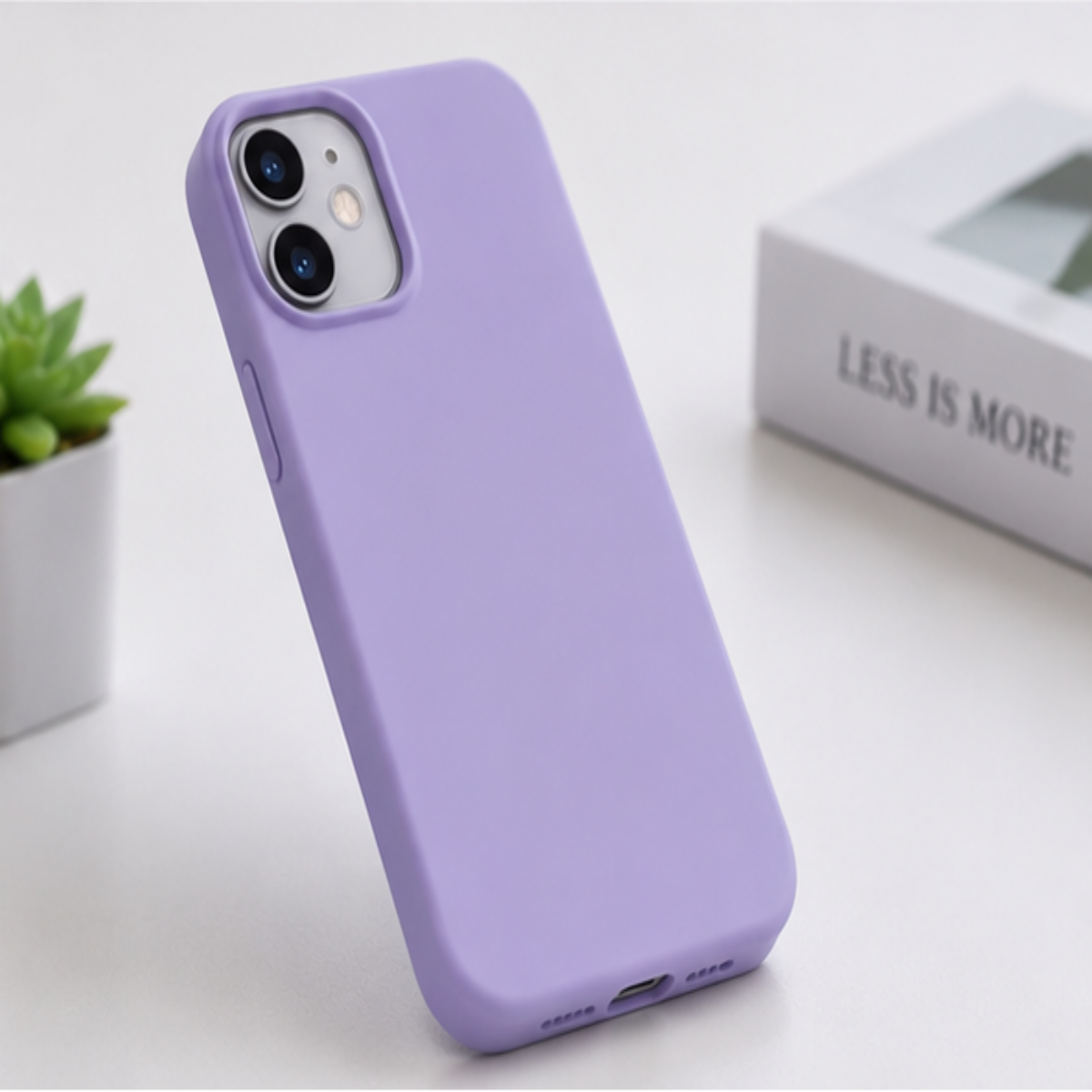 A lavender purple phone case shown standing upright on a white tabletop, with a small plant and a book in the background.